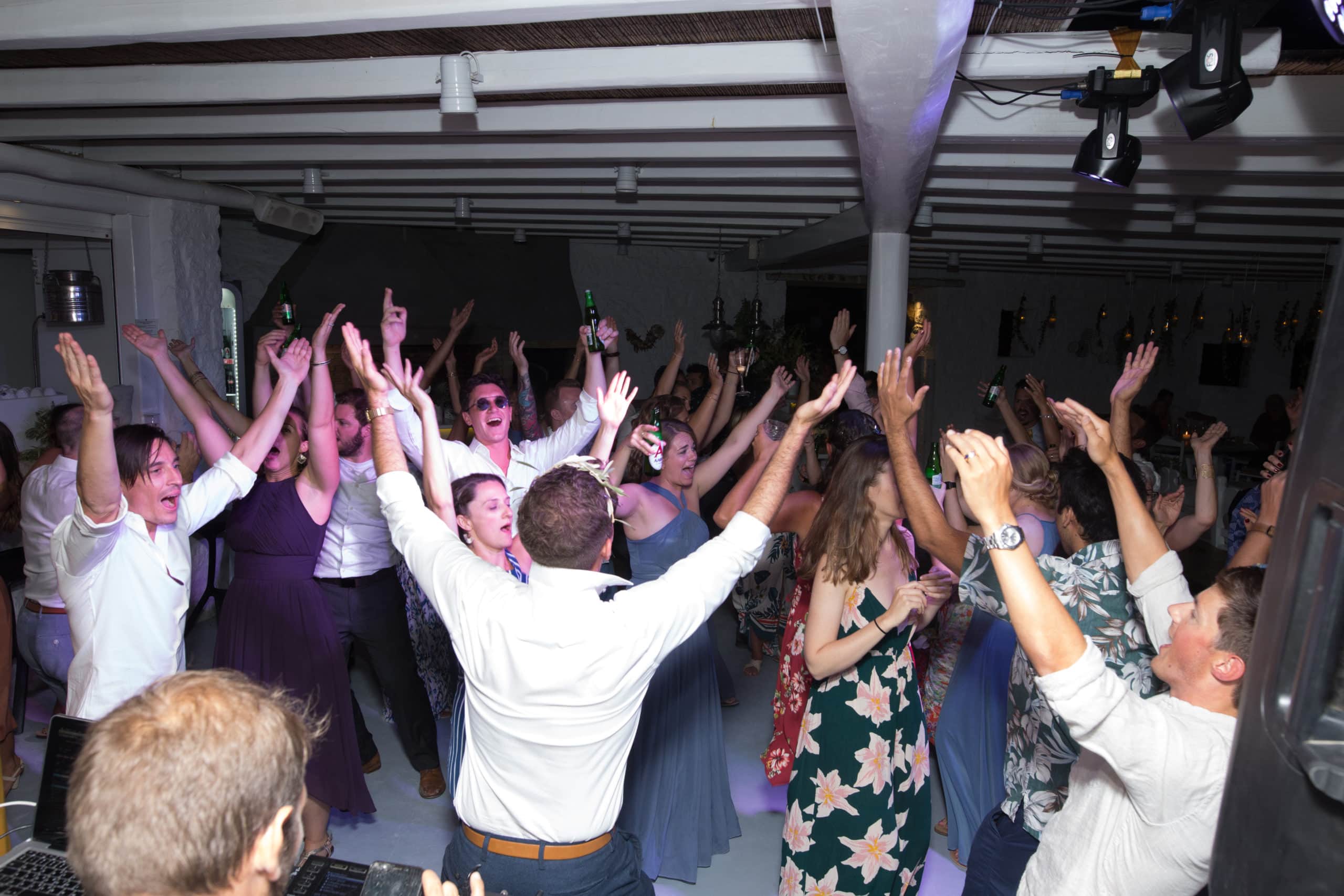 Wedding guests with their hands in the air on a packed dance floor under moving-head lights in Mykonos