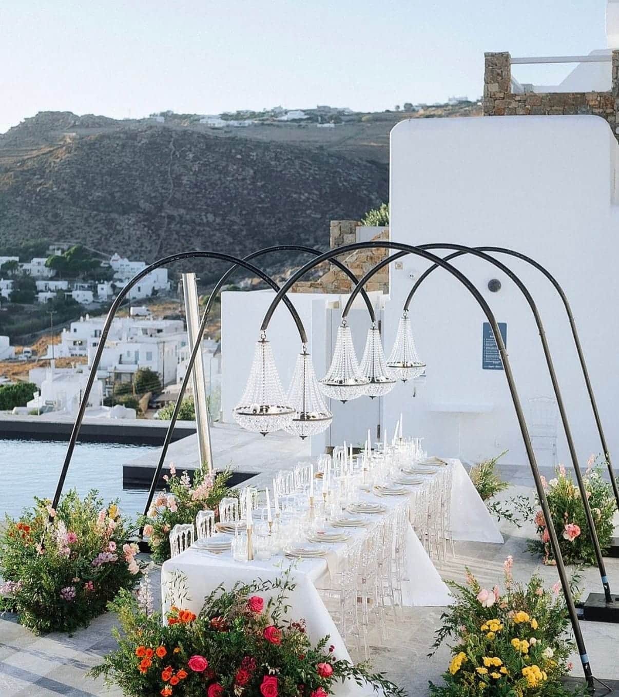 Long white dinner table under metal arches on a terrace overlooking whitewashed Cycladic houses
