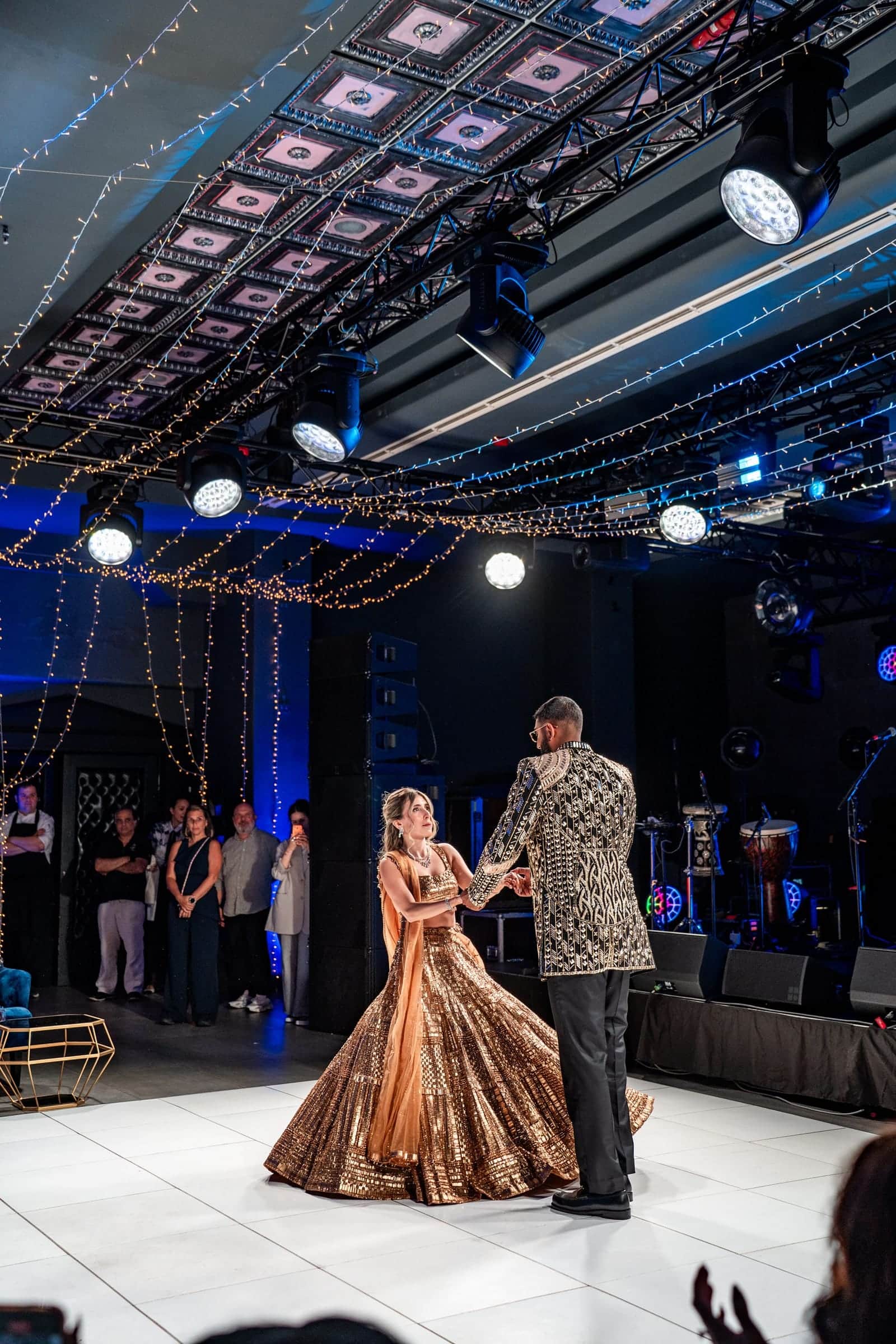 Bride and groom dancing on a white dance floor, watched by guests, beneath a lighting truss with moving heads and festoon lights