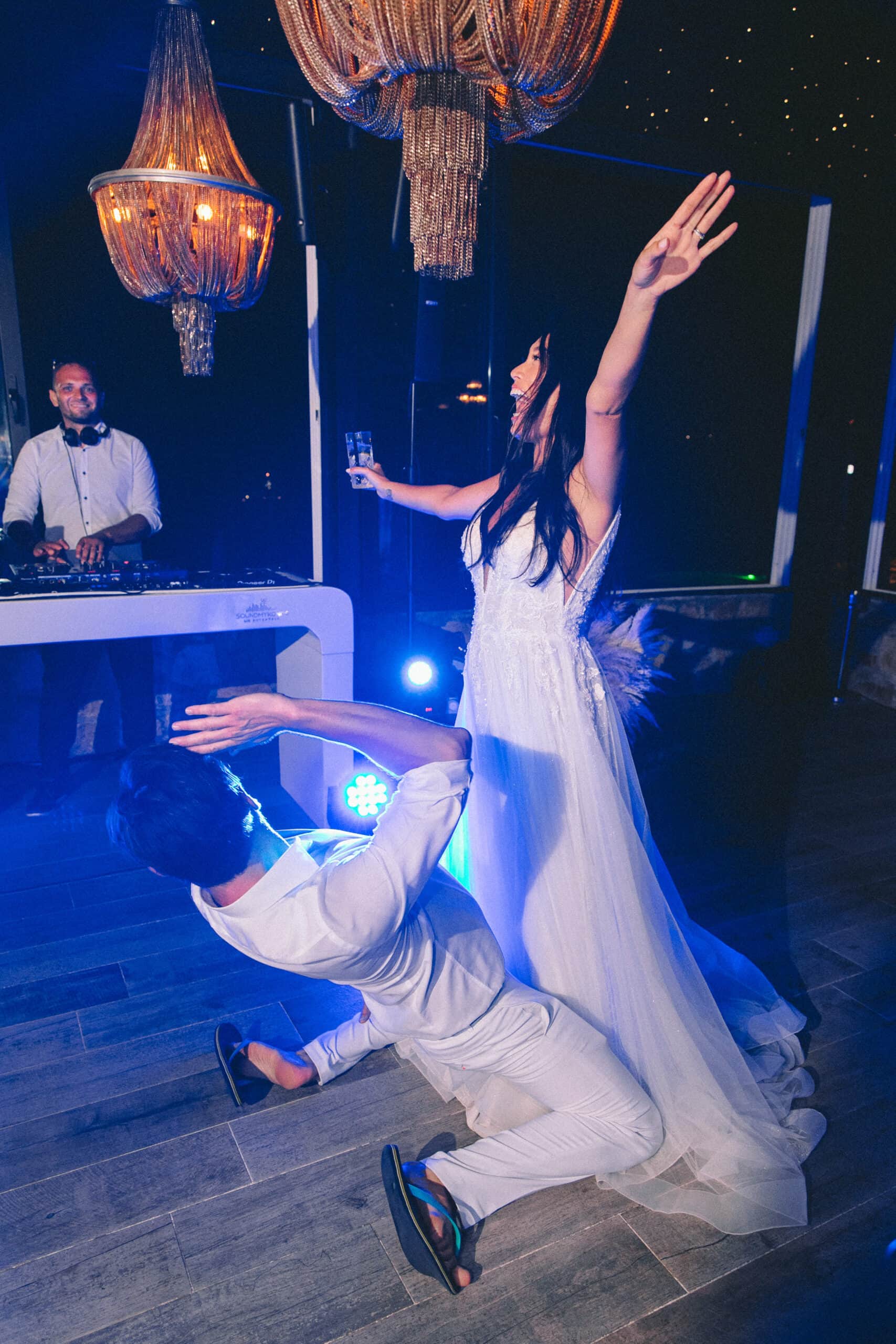 Groom drops to the floor in front of the bride on a blue-lit dance floor, DJ at the booth behind