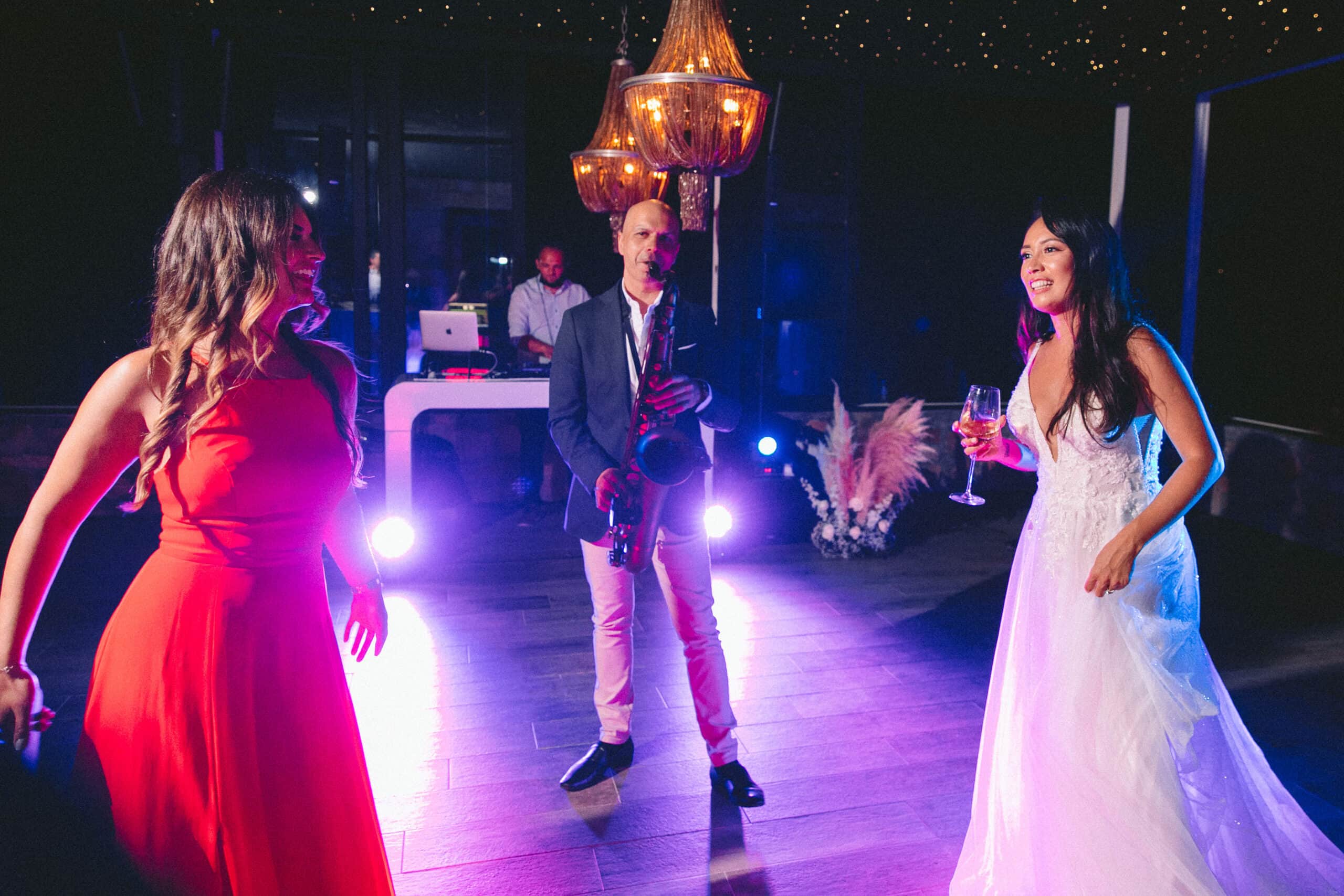 Live saxophonist playing between the bride and a guest on a lit dance floor, DJ booth behind