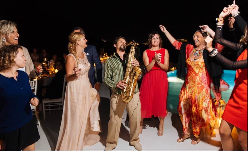 Saxophonist playing among dancing guests beside a pool at night