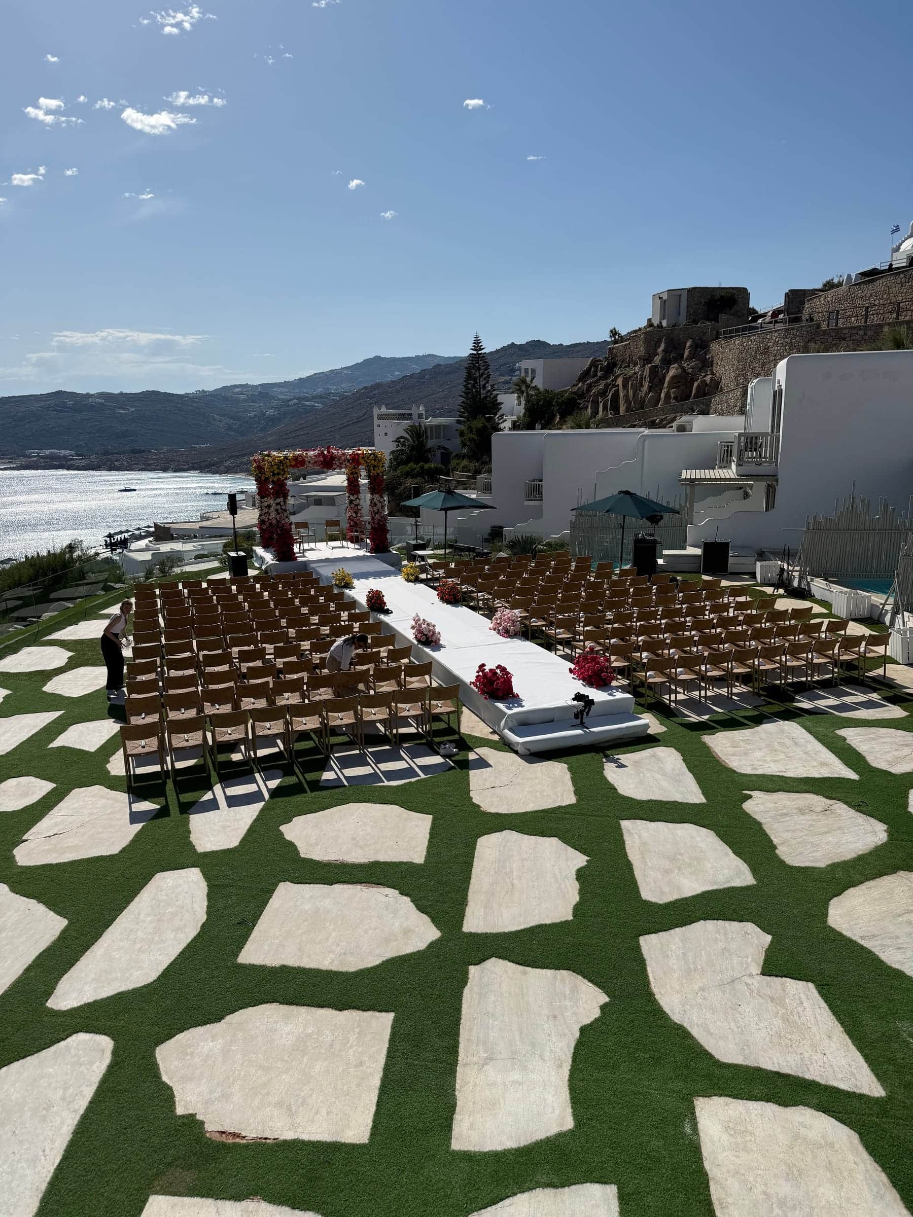 Seaside lawn in Mykonos set for a wedding ceremony with rows of chairs, a white aisle and a floral canopy
