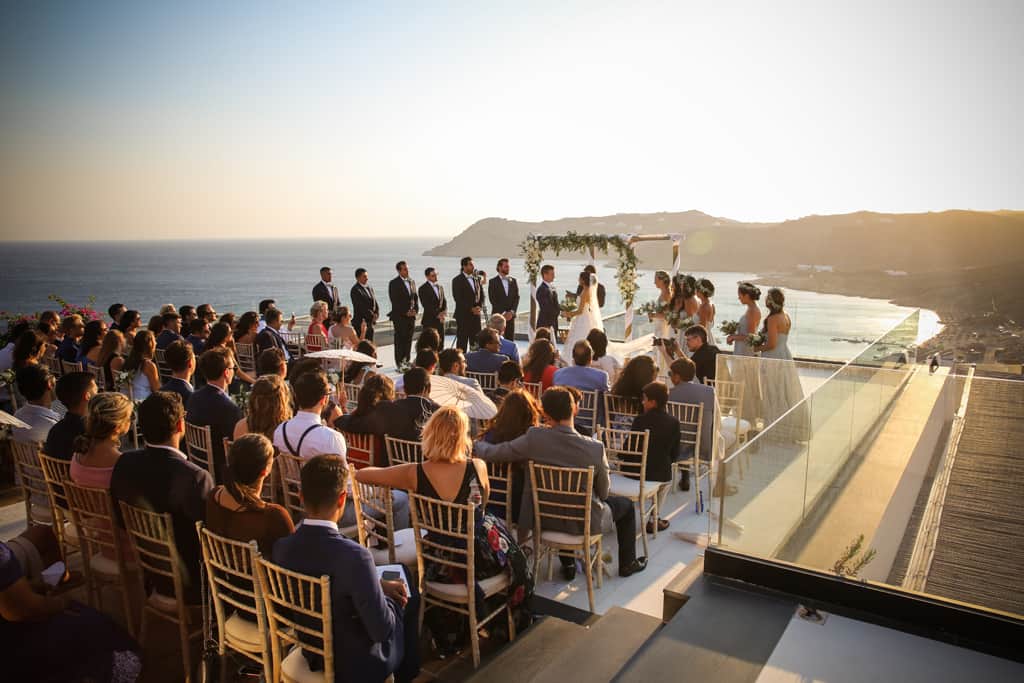 Sunset wedding ceremony on a clifftop terrace, guests seated facing the couple and the Aegean