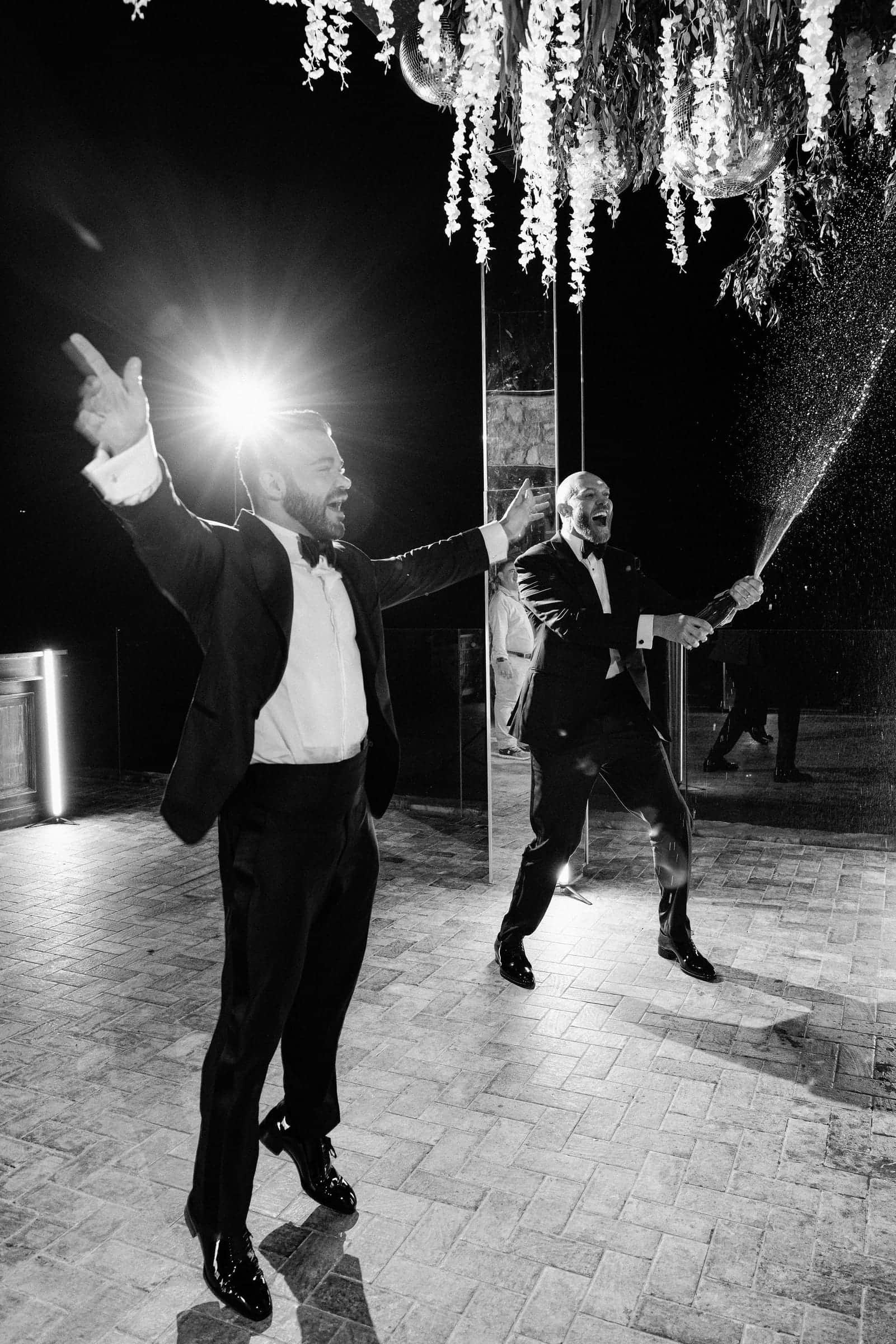 Black-and-white photo of a man in a tuxedo dancing as a guest sprays champagne