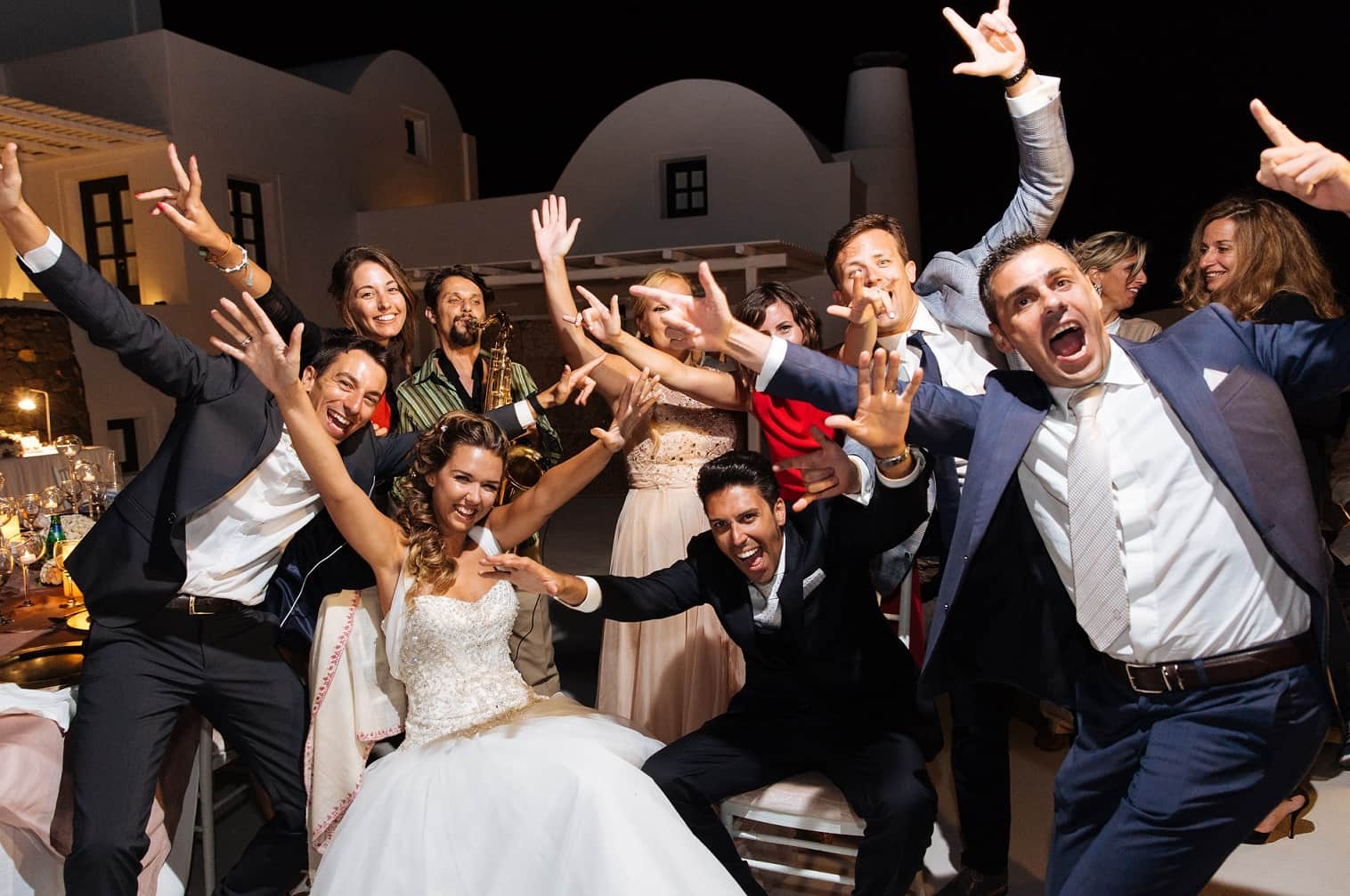 Bride, groom and guests celebrating with arms raised in front of whitewashed Cycladic buildings