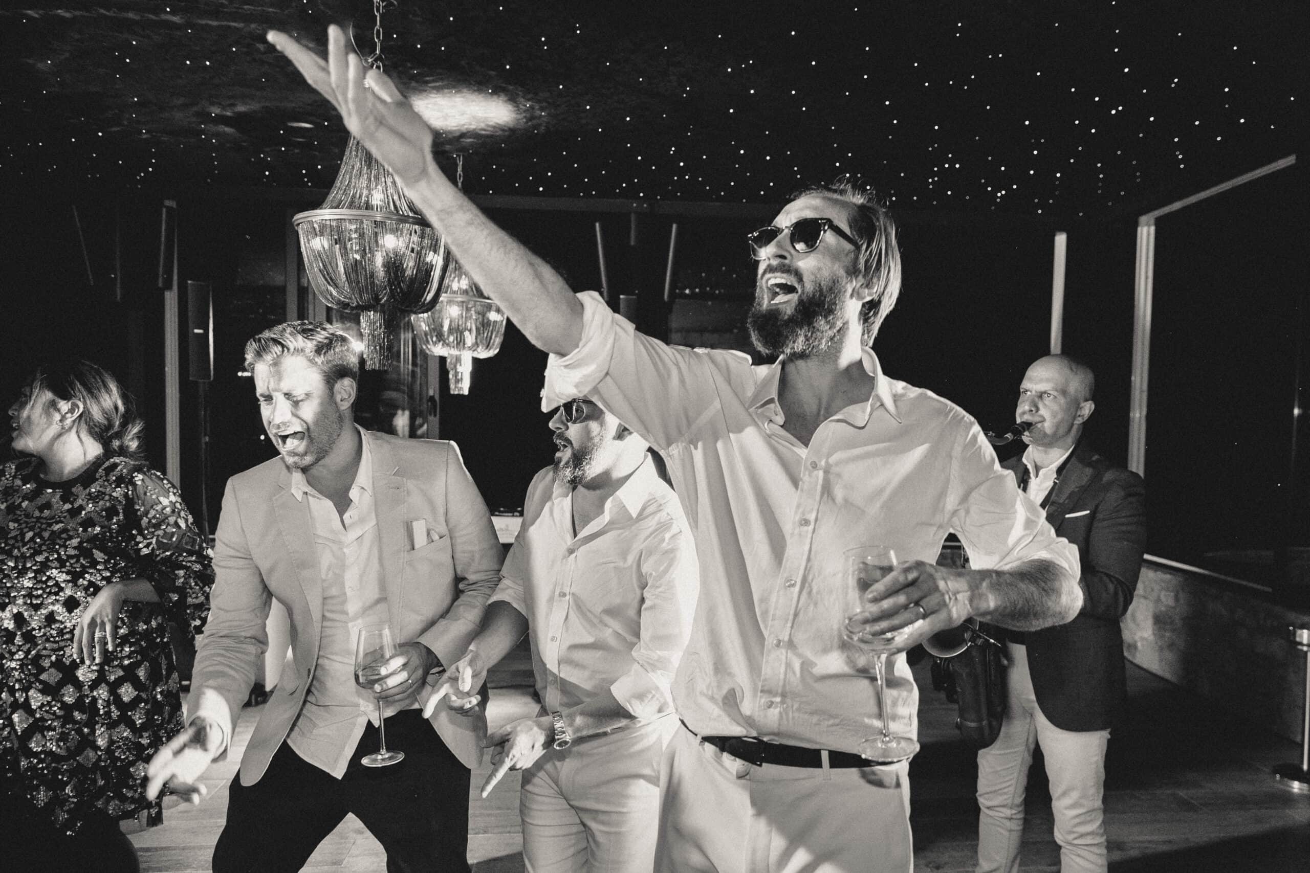 Black-and-white photo of wedding guests singing along as a saxophonist plays